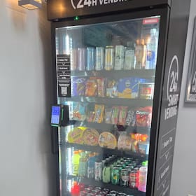 Branded 24 hour smart vending cooler with a lit glass front and a side wrap panel, stocked with snacks and drinks