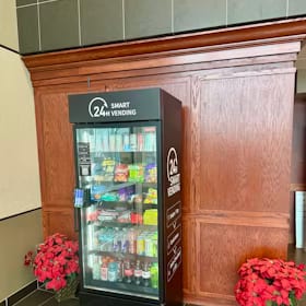 Smart vending cooler with a 24 hour header panel set against wood paneling in a hotel lobby, flanked by poinsettias