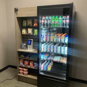 Small micro market corner with a glass-front drink cooler beside open wood shelving of snacks and a checkout screen